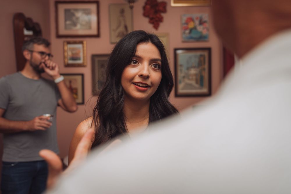 Young woman smiling at a social gathering with framed artwork on pink walls and guests in background.