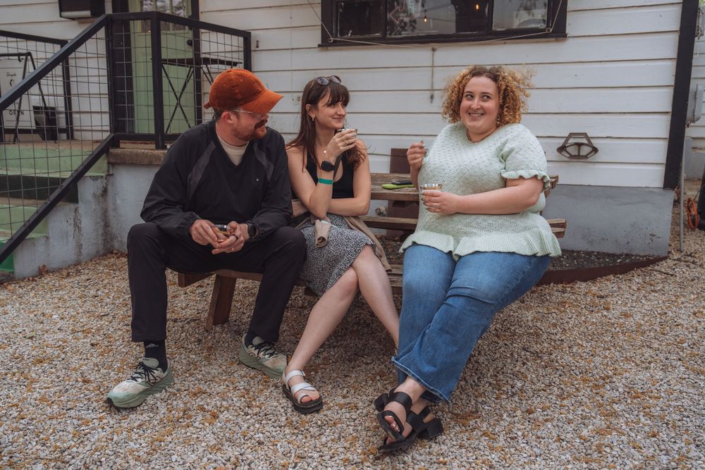 Three friends laughing and socializing on a wooden bench outside a white building on a gravel patio.