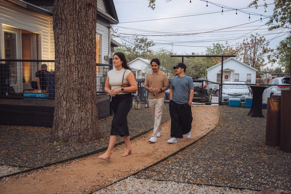 Three people walking along a garden path at an outdoor evening gathering near a house with string lights.