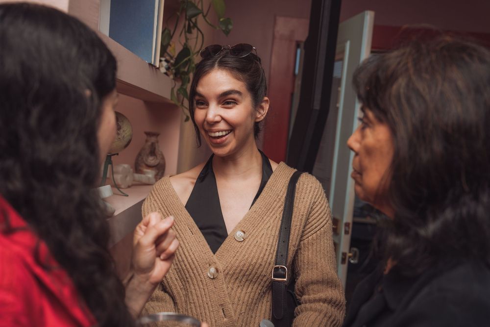 Three women engaged in lively conversation at an indoor social gathering, smiling warmly in a cozy setting.