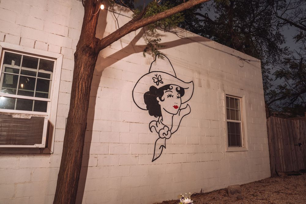 Vintage cowgirl mural painted on white brick building exterior at night with tree in foreground.