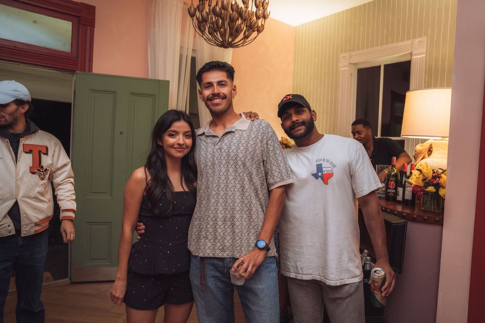 Three friends smiling together indoors at a casual social event beneath a decorative chandelier.