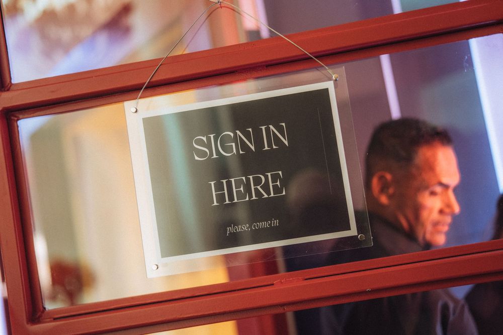 A reflective sign reading Sign In Here please come in hanging in a red-framed window with a man visible in background.