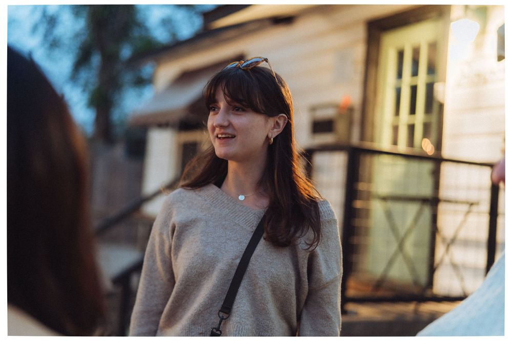 Young woman with dark hair smiling outdoors near a house, wearing a gray sweater and crossbody bag.