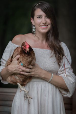 Smiling brunette woman in white off-shoulder dress holding a brown hen while posing outdoors.