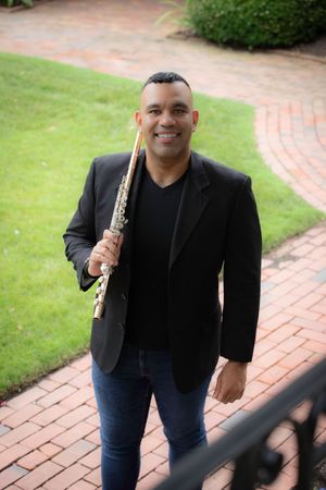 Smiling male flutist in black blazer holding flute on brick garden pathway.