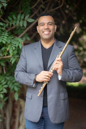 Smiling male flutist in gray blazer holding gold flute outdoors among green foliage.