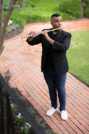 Male musician playing flute on brick garden path surrounded by greenery.