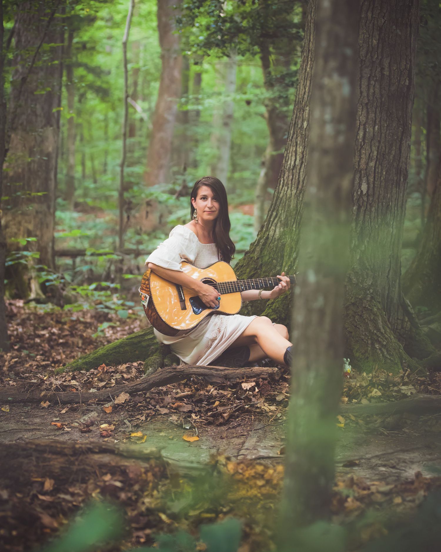 Young woman in white dress sitting against tree playing acoustic guitar in lush green forest setting.