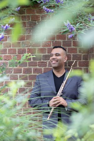 A smiling man holds a flute while posing against a brick wall surrounded by green foliage.