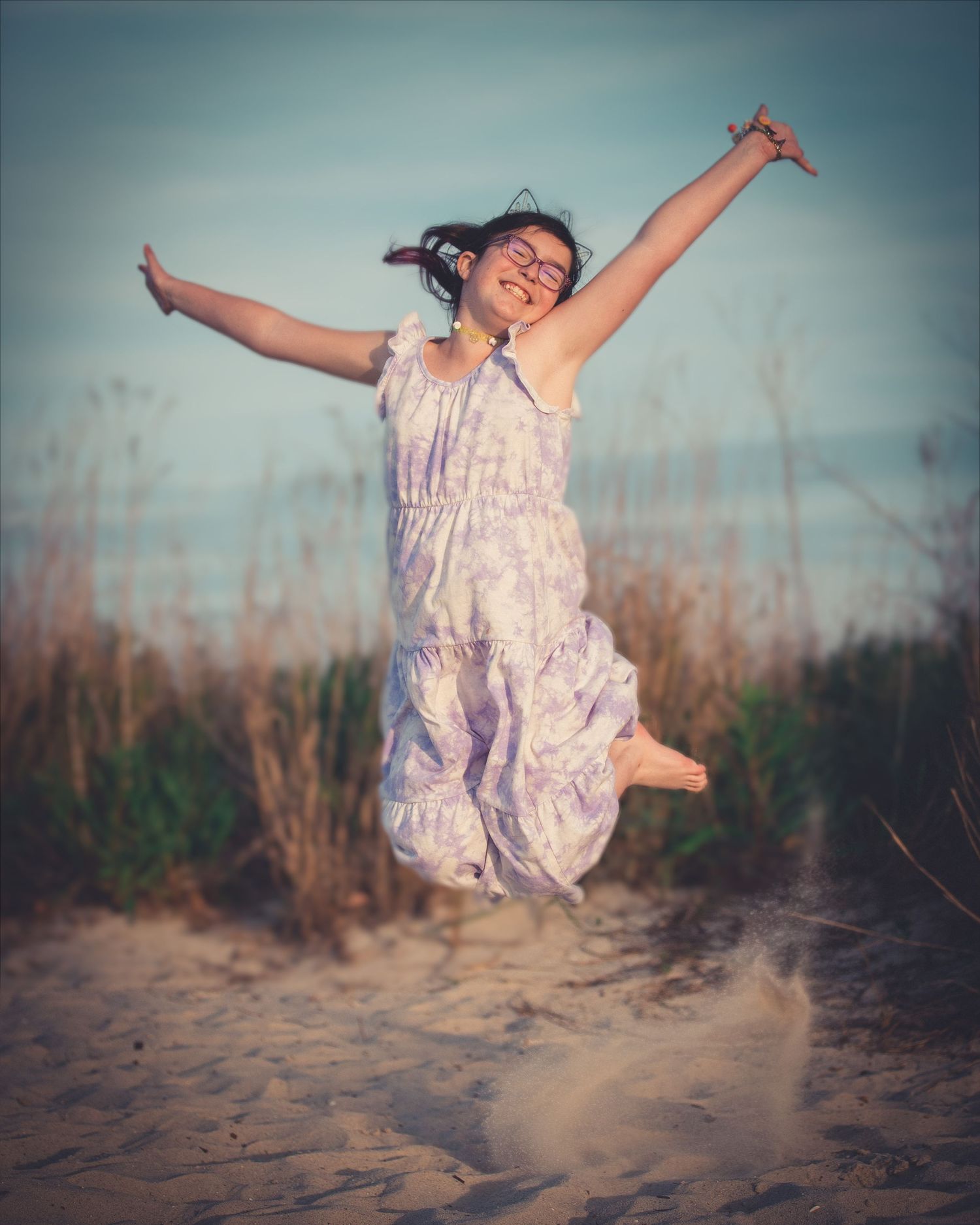 A young girl in a floral dress joyfully jumps with arms wide open on a sandy beach at sunset.