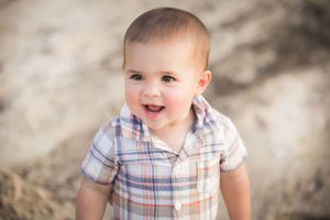 Happy baby boy in plaid shirt smiling outdoors on sandy background.