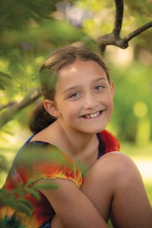 Smiling young girl in tie-dye shirt sitting under green tree branches outdoors.