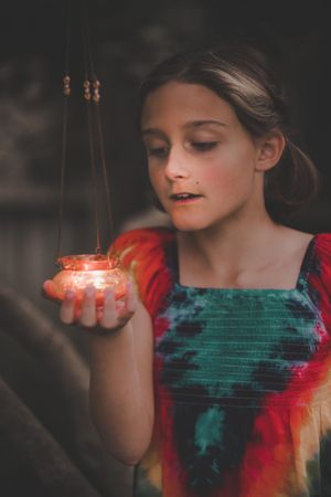 Young girl in tie-dye dress holding glowing red candle lantern in dim light.