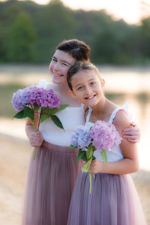 Two smiling girls in white tops and purple tulle skirts holding hydrangea bouquets.
