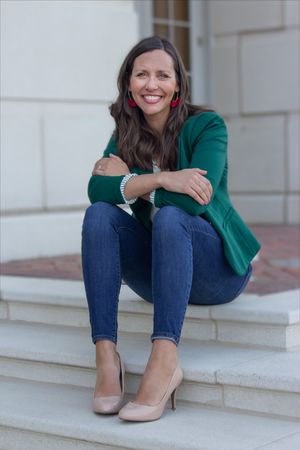 Smiling woman in green blazer and jeans sitting on white stone steps outdoors.
