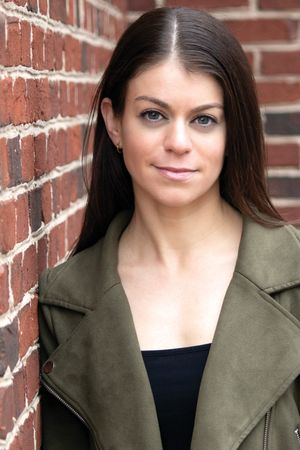 Young woman with dark hair in olive green jacket leaning against red brick wall.
