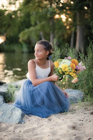 Young girl in blue tulle skirt holding colorful flower bouquet by a lake.