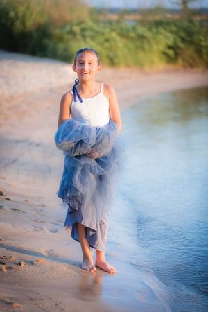 Smiling girl in white top and blue tulle skirt walking barefoot on sandy beach.