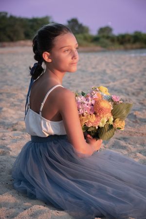 A teenage girl in a blue tulle skirt sits on a sandy beach holding a colorful bouquet at sunset.