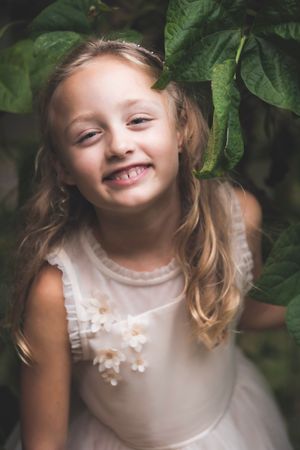 A smiling young girl in a white dress peeks through large green leaves in a garden setting.