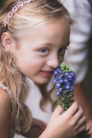 Smiling blonde girl with headband smelling purple wildflower bouquet outdoors.