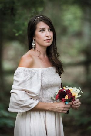 Woman in white off-shoulder dress holding red and yellow bouquet in forest setting.