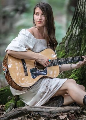 A woman in a white dress sits against a mossy tree in a forest playing an acoustic guitar.