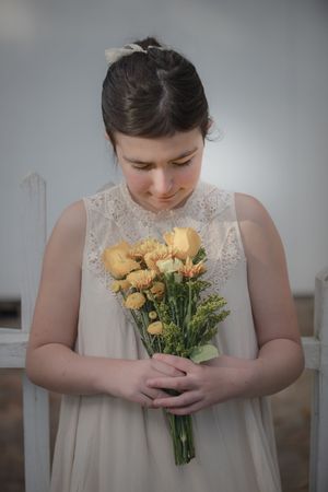 Young girl in cream lace dress holding yellow flower bouquet, looking down.