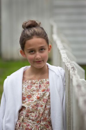 Young girl with bun hairstyle in floral dress and white jacket leaning on white fence.