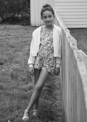 Young girl in floral romper and white jacket leaning against wooden fence, black and white photo.