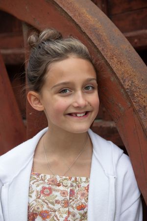 Young girl with bun hairstyle smiling beside rusty vintage wheel outdoors.