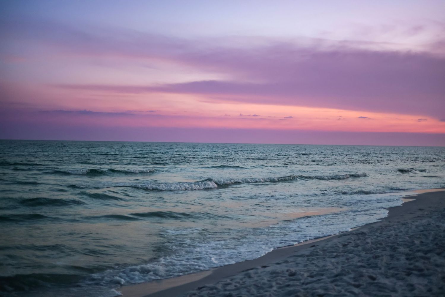 A stunning pink and purple sunset over calm ocean waves on a quiet sandy beach shoreline.