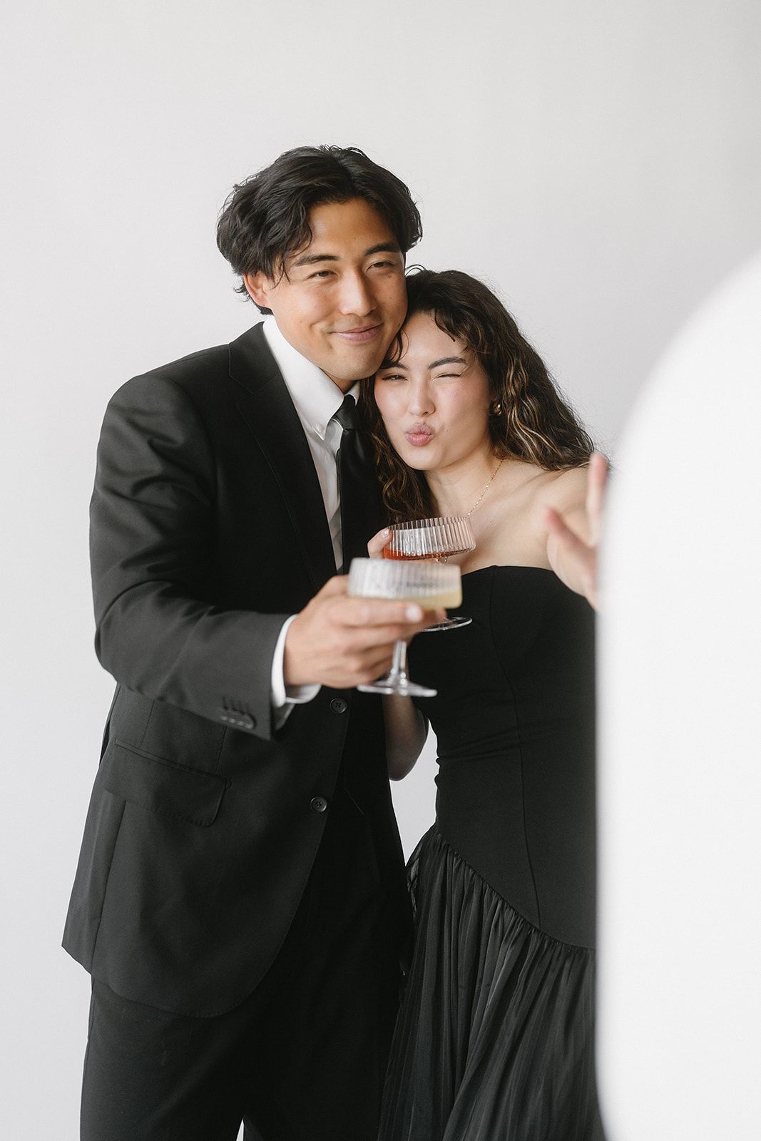 Couple in black formal wear posing with champagne glass, woman making kissy face, white background.