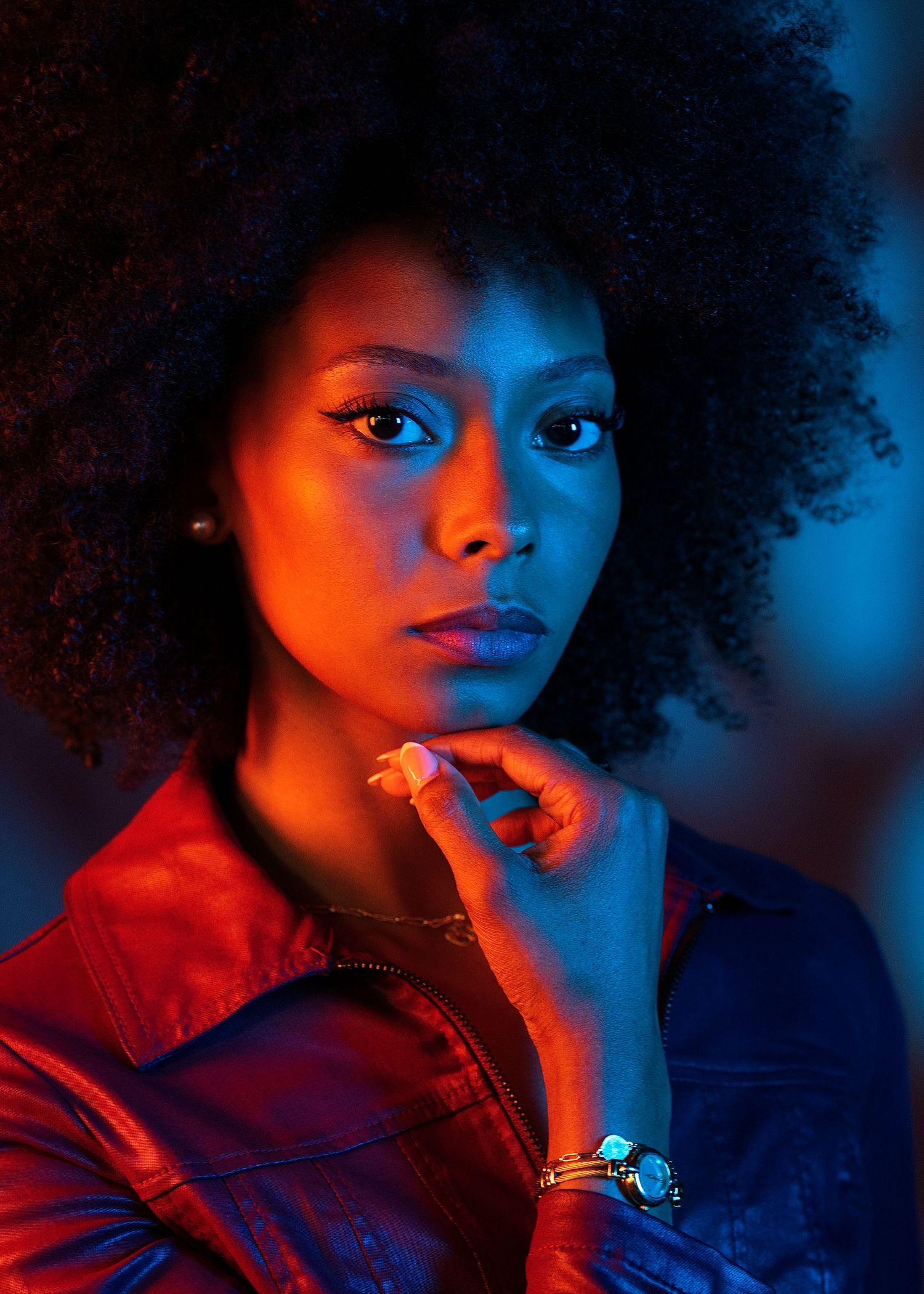 Close-up portrait of young Black woman with afro hair in red jacket under dramatic orange and blue neon light.