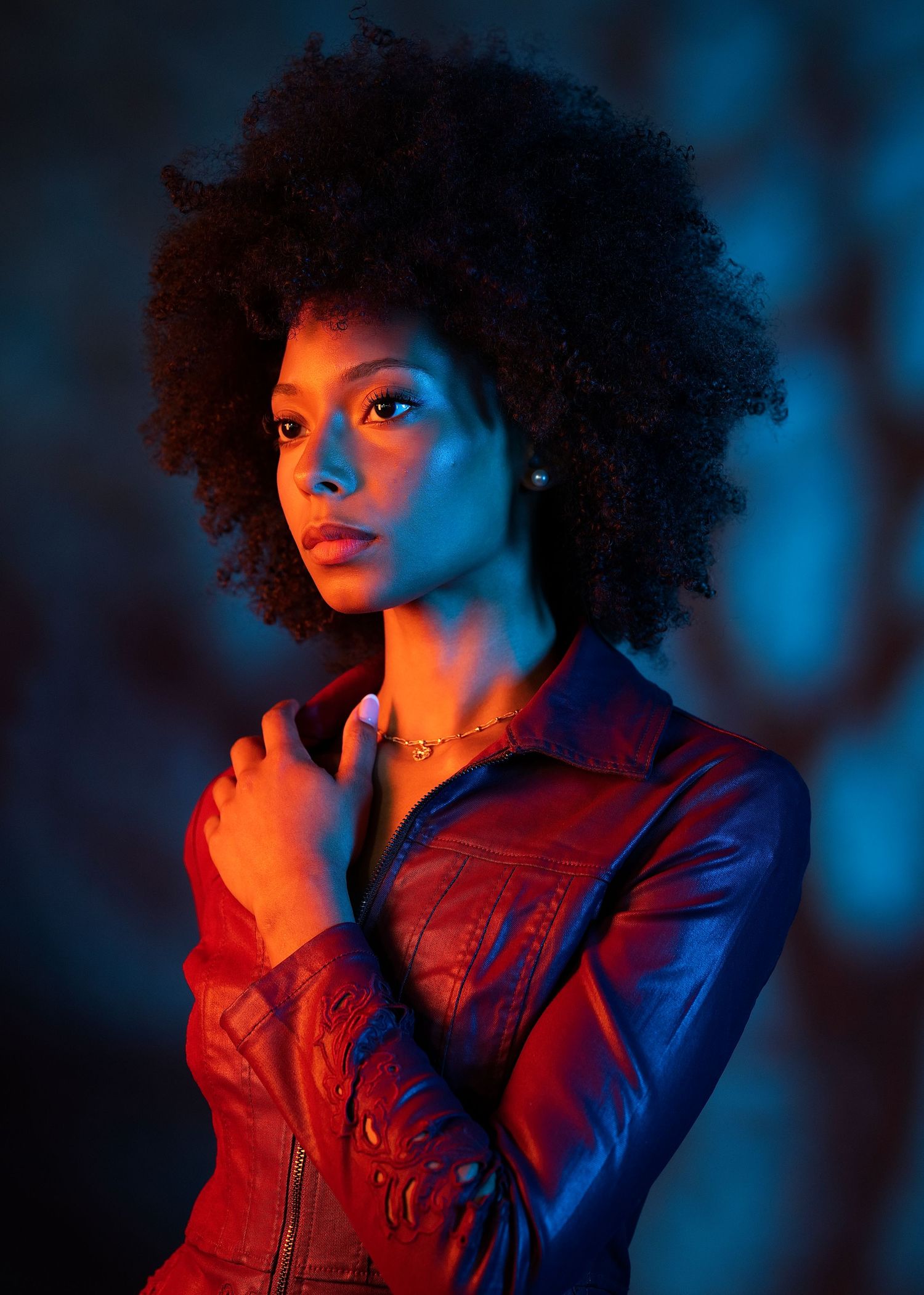 Young Black woman with natural afro in red leather jacket posed under moody blue and orange studio lighting.