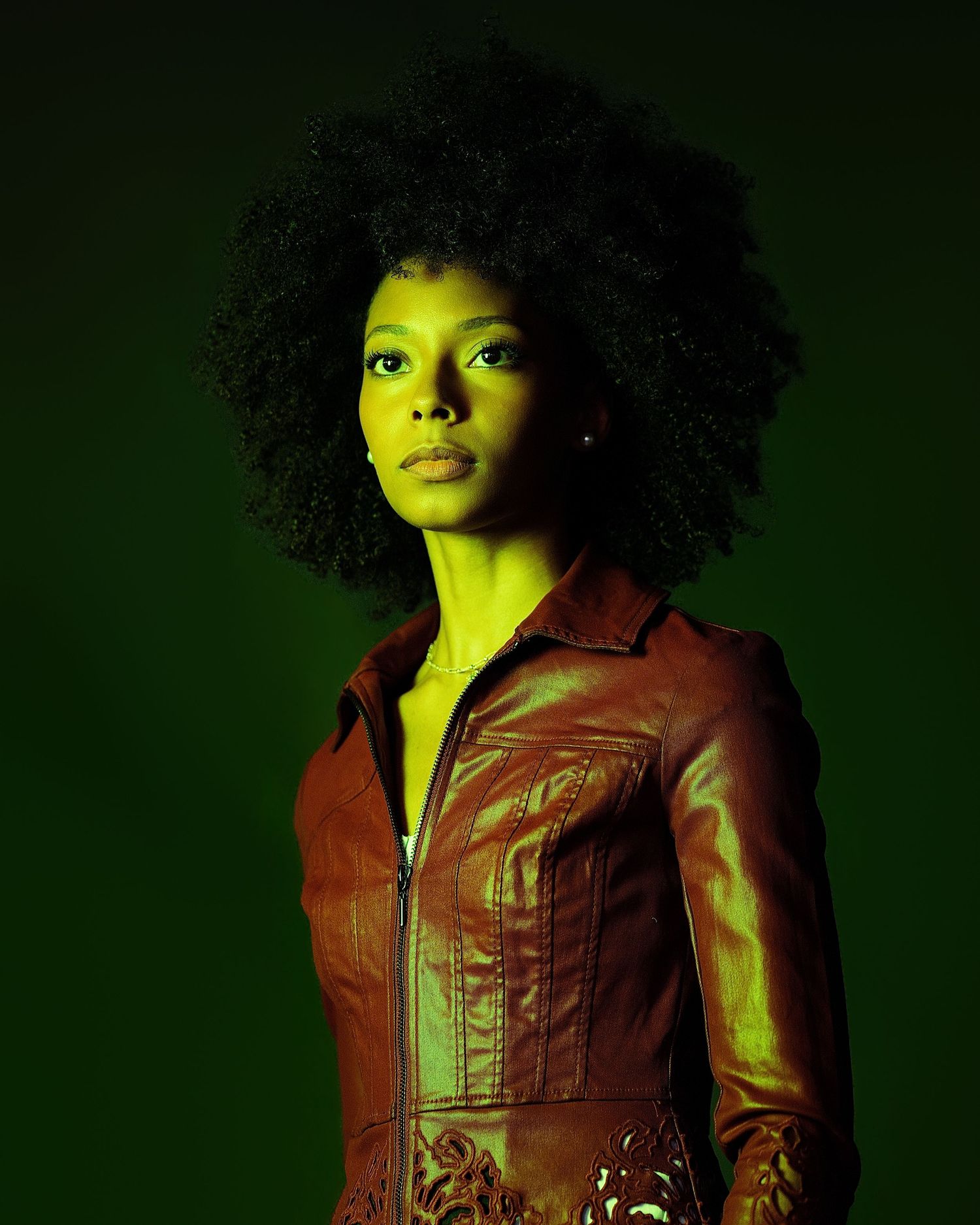 Woman with natural afro hair wearing brown leather jacket poses under striking green studio lighting.