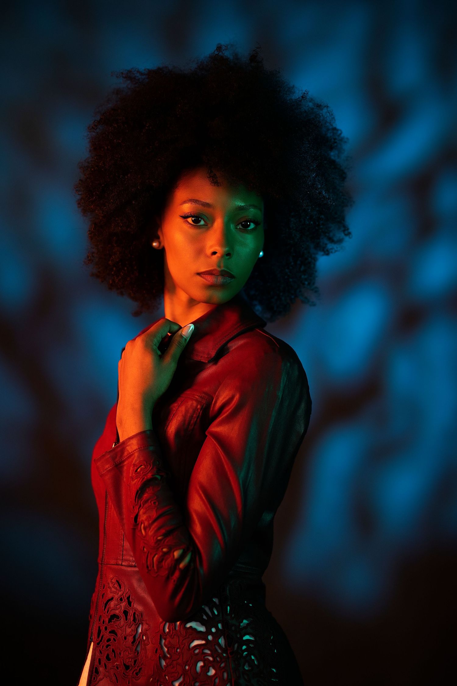 Young Black woman with afro hair in red leather jacket posed under dramatic blue and orange neon lighting.