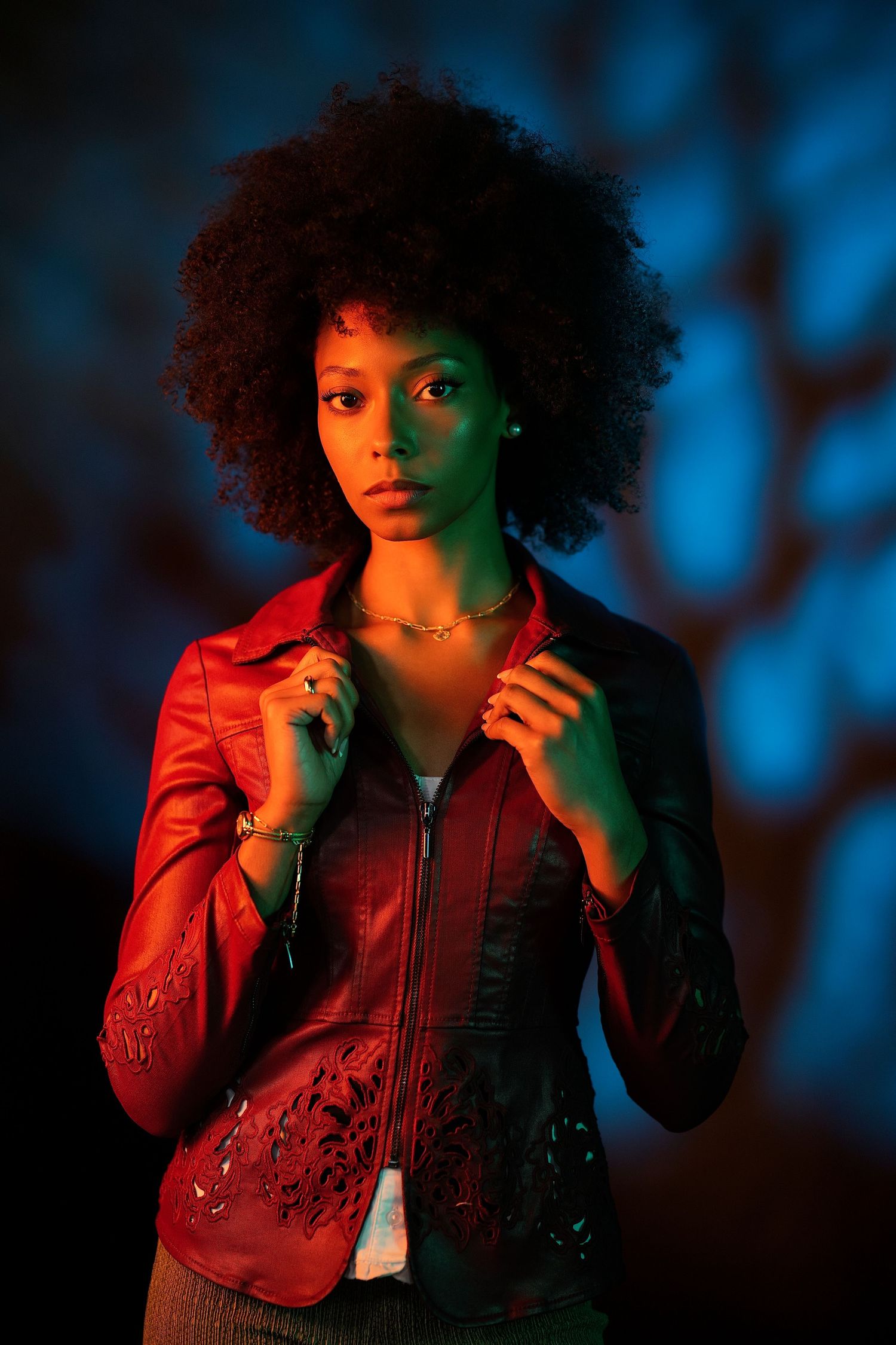 Young Black woman with natural afro in red leather jacket posing under colorful neon lights with tree shadow.