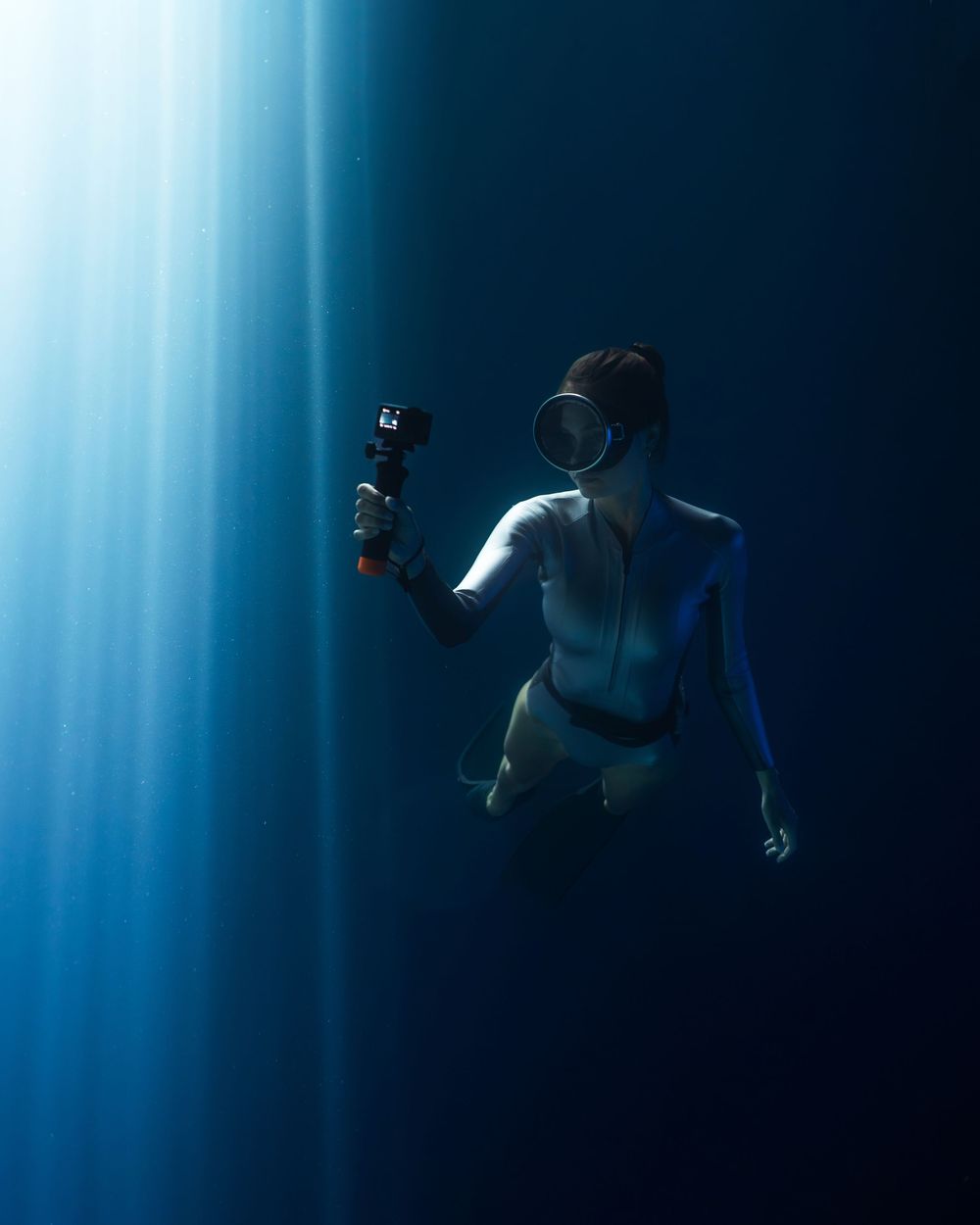 Underwater diver in white wetsuit holding an action camera, illuminated by dramatic blue light rays in deep ocean.