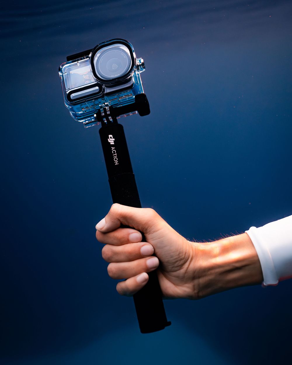 Hand holding a DJI Action camera in waterproof housing on a selfie stick underwater with blue ocean background.