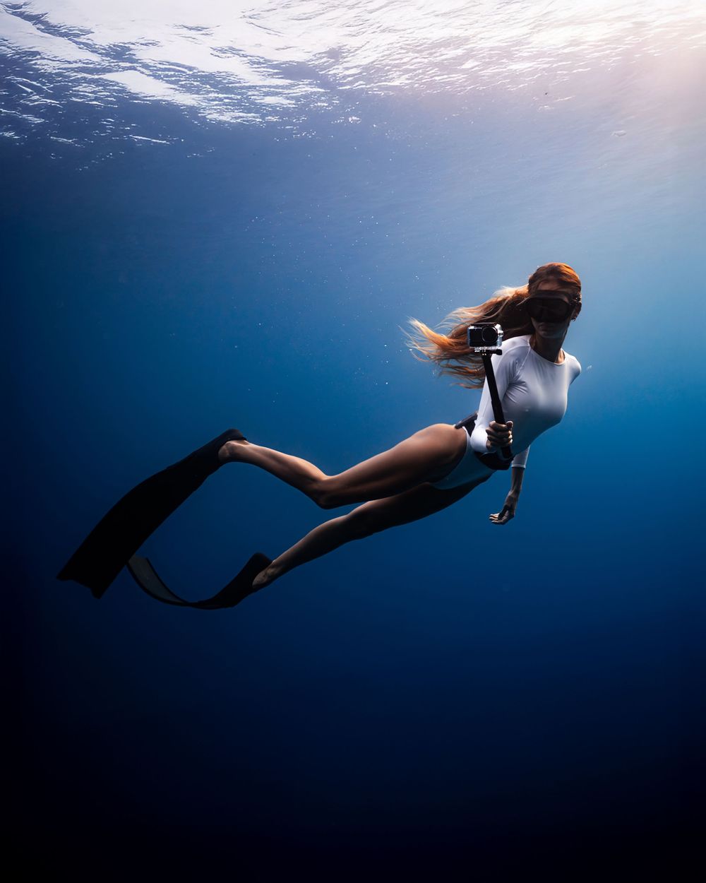 A female freediver in a white wetsuit glides gracefully underwater in deep blue ocean, holding a camera with black fins.