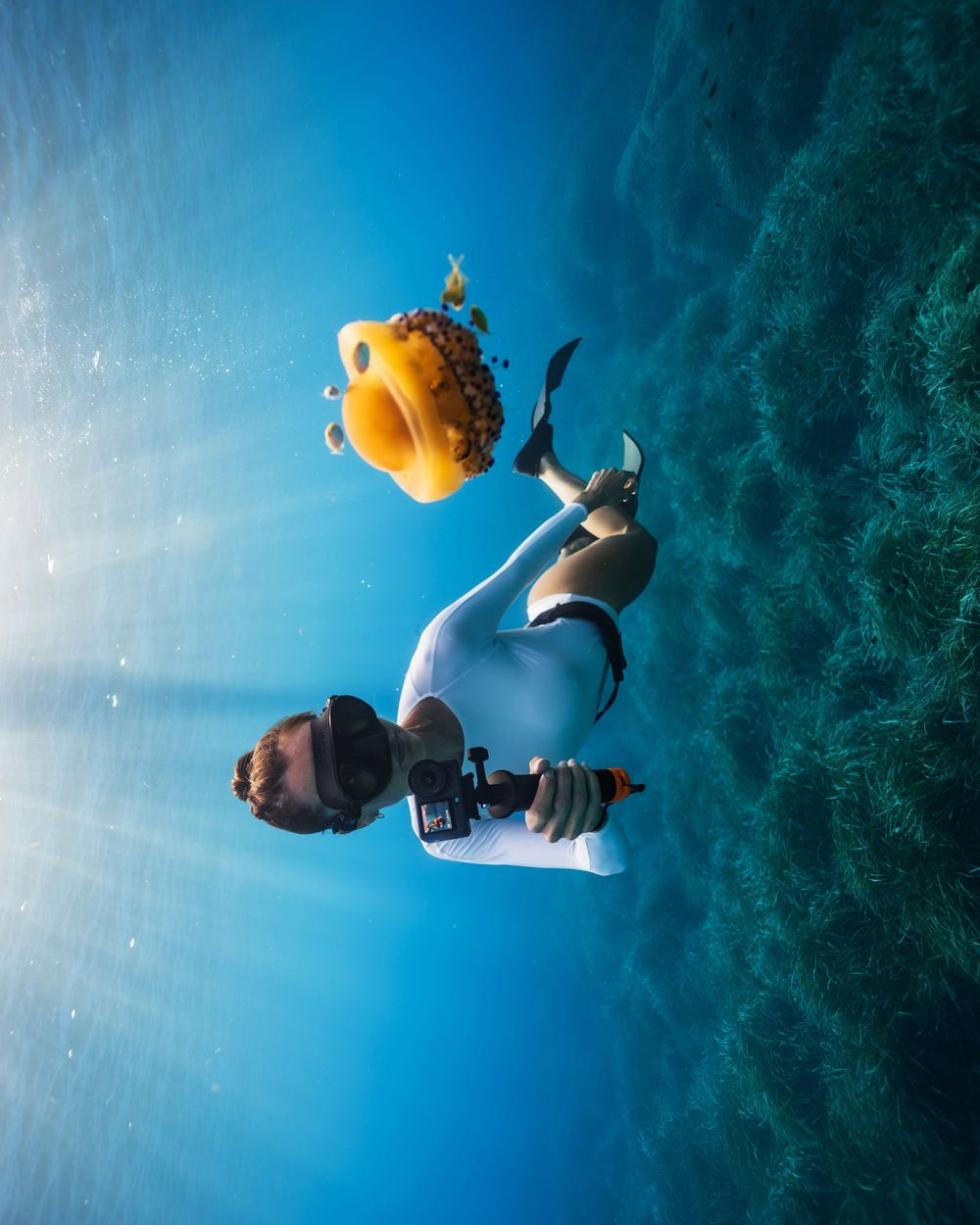Underwater freediver in white wetsuit holding a camera, swimming near coral reef with a yellow fish float above.