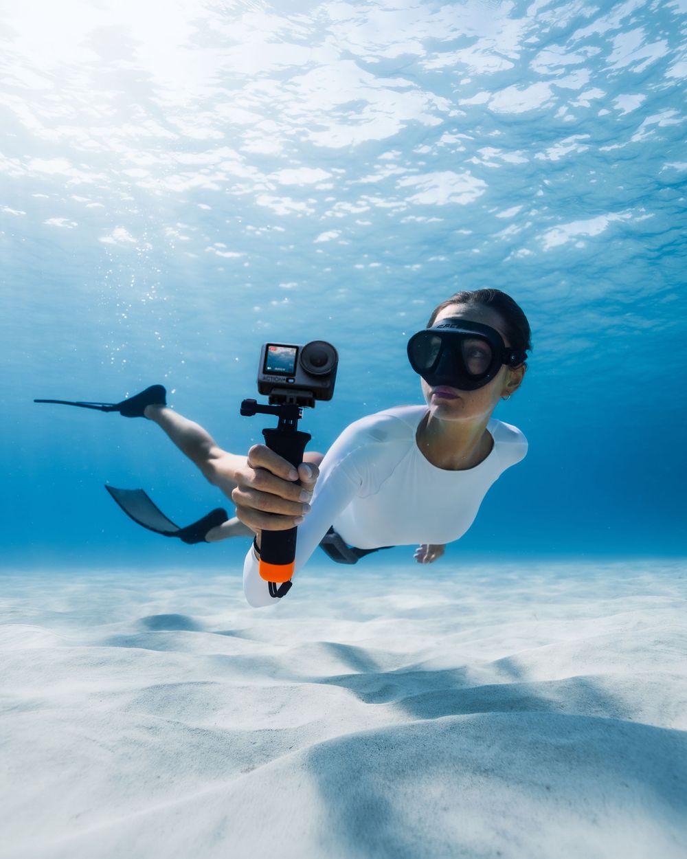 Freediver underwater holding a GoPro action camera on a pole, wearing a white wetsuit and diving mask in clear blue ocean.