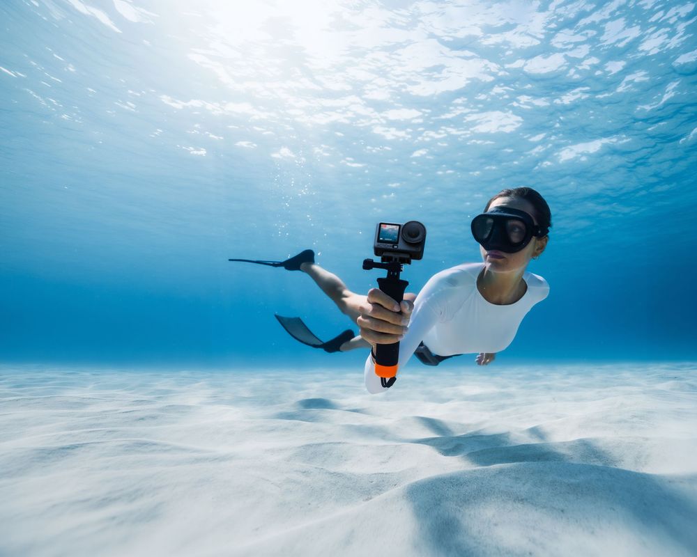 Snorkeler diving underwater holding a GoPro camera on a float, wearing fins and mask in clear blue tropical ocean.