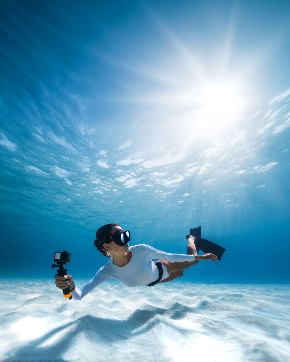 Freediver in white wetsuit glides underwater holding a GoPro camera, with sunlight streaming through crystal clear blue ocean.