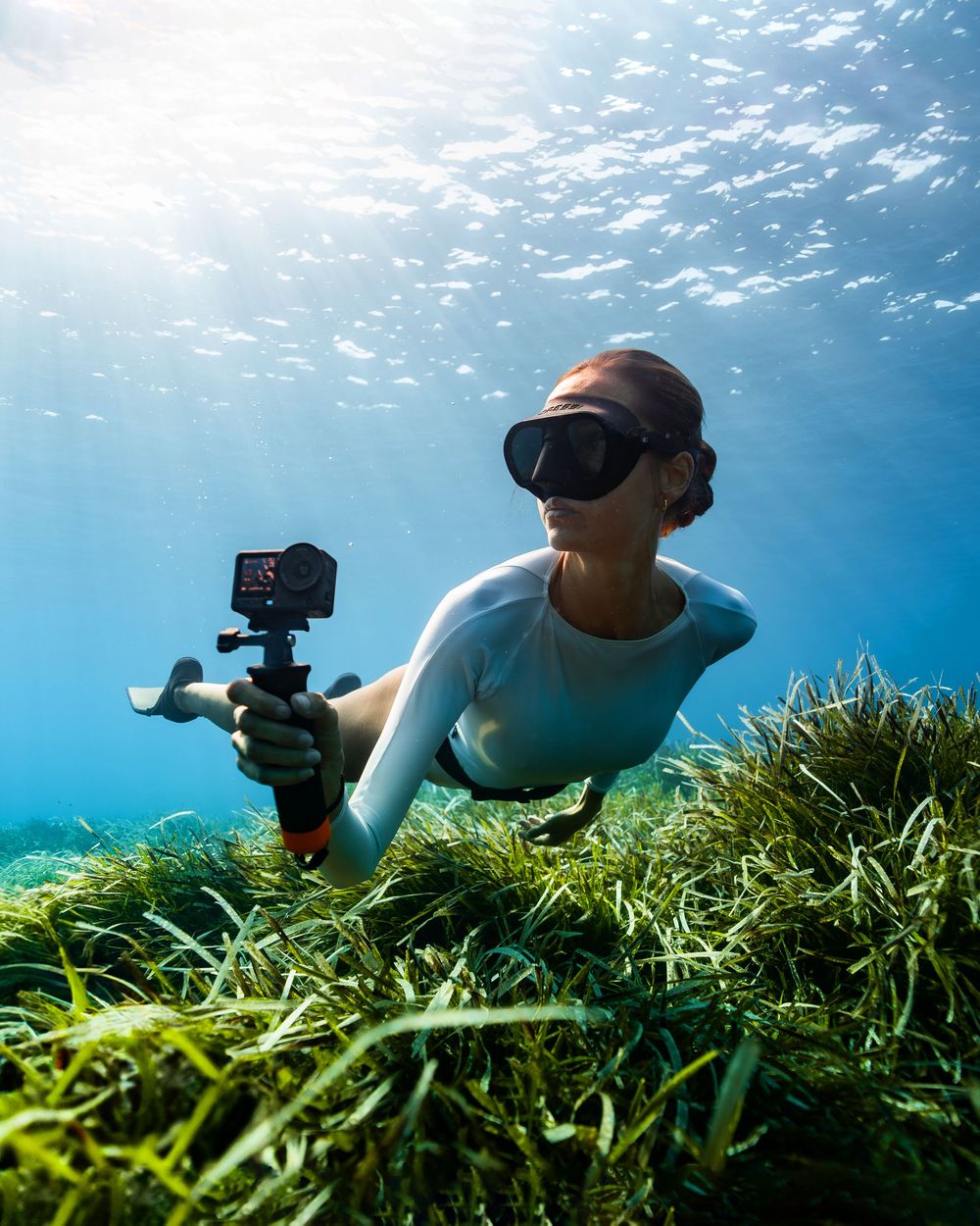 Woman in diving mask holds action camera while freediving over lush seagrass in clear blue ocean water.