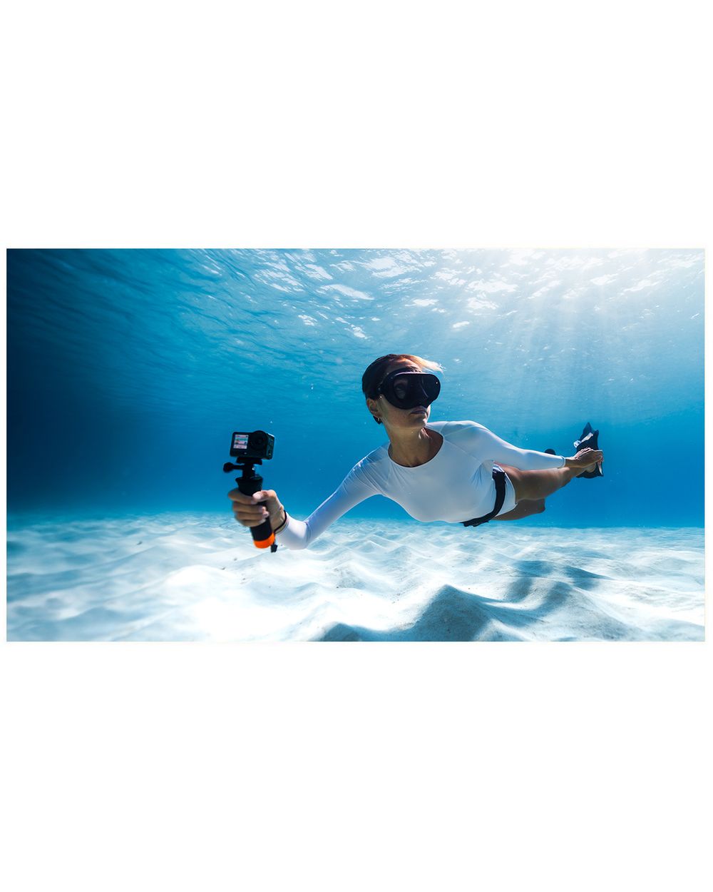 Person swimming underwater with a GoPro camera on an orange handle, wearing a white wetsuit and diving mask.