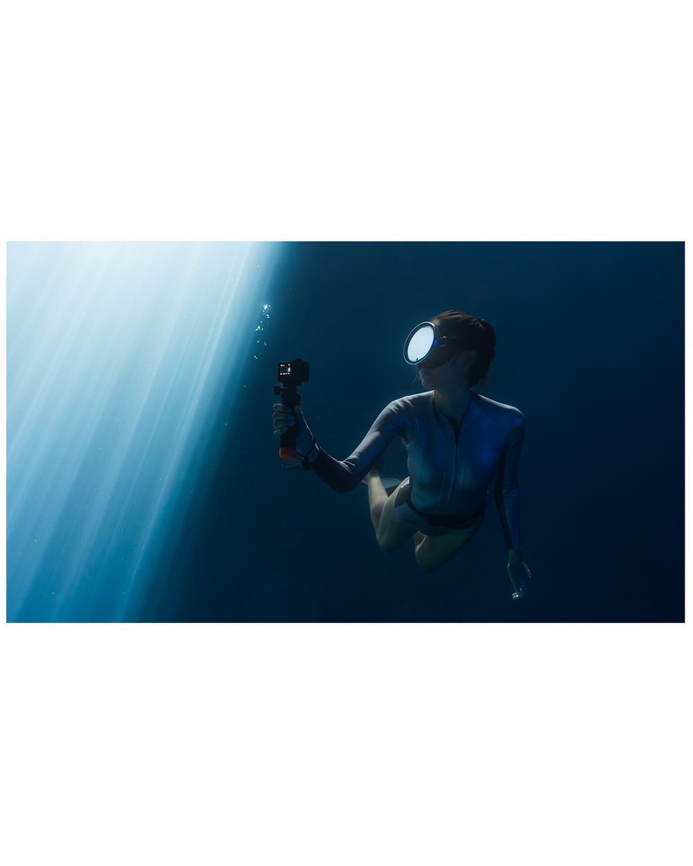 Underwater photographer in diving suit capturing footage with a camera, illuminated by light rays in deep blue ocean.