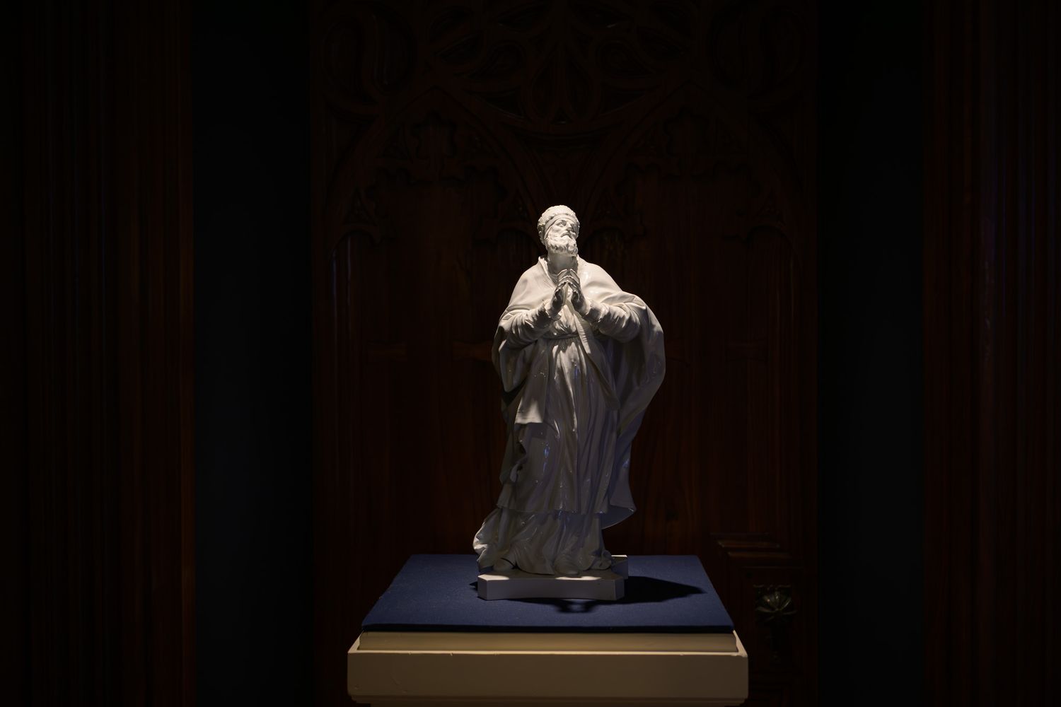 A small ceramic figurine of a robed religious figure displayed on a pedestal in a dimly lit museum exhibit.